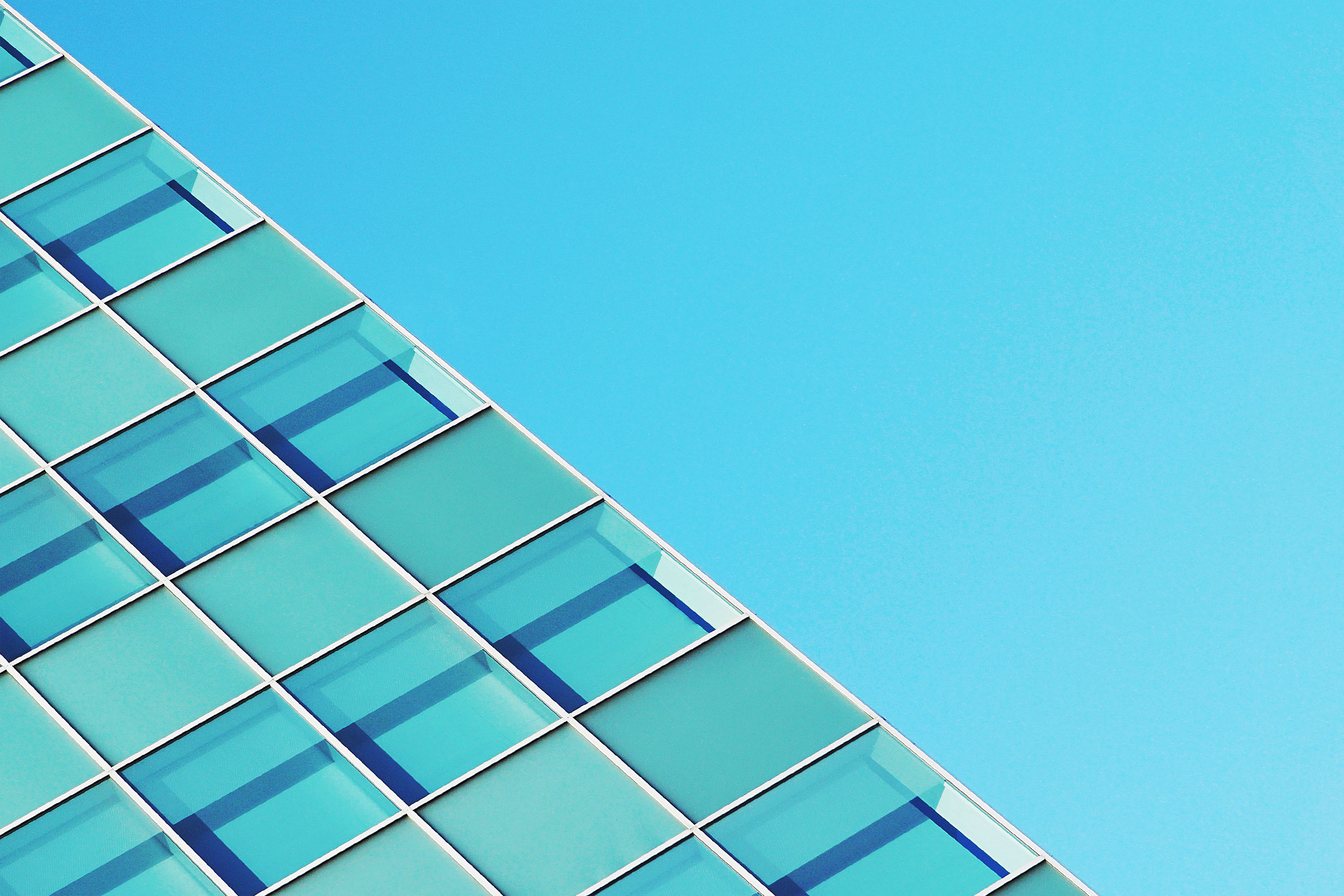 A modern glass building facade with teal-blue reflective windows stretching upward against a clear blue sky, conveying a clean and fresh urban atmosphere